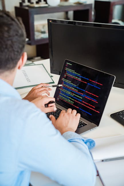 A person working at a desk with a laptop displaying lines of colourful code, surrounded by a computer monitor, a notebook, and a pen. The workspace appears organized with a modern aesthetic, featuring